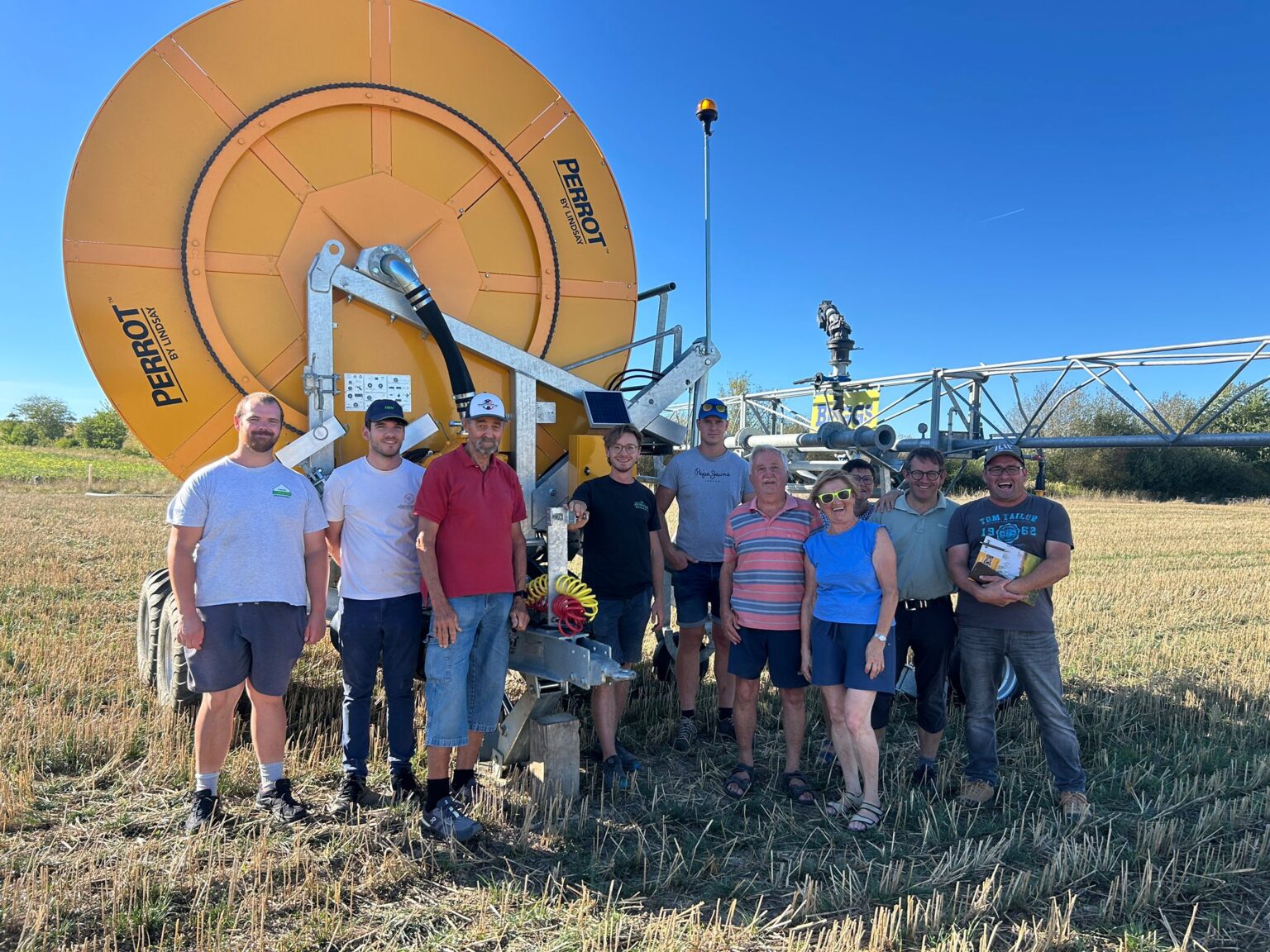Eine Gruppe von zehn Personen steht lächelnd vor einer großen Bewässerungsmaschine auf einem sonnigen, abgeernteten Feld, im Hintergrund sind der blaue Himmel und landwirtschaftliche Maschinen zu sehen.